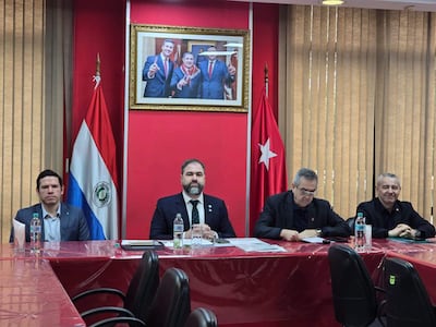 Rodrigo Nicora y otros diputados en traje negro sentados en mesa con banderas de Paraguay y Turquía, revisando documentos.