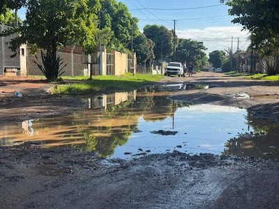 Dos personas de pie conversan al lado de un charco en una calle con baches y vegetación, con un auto estacionado al fondo.