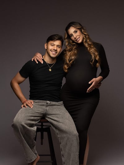 Ángel Romero en camiseta negra y pantalones claros, abrazando a su pareja embarazada vestida de negro, ambos sonrientes en un entorno neutro.