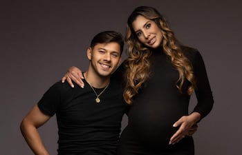 Ángel Romero en camiseta negra y pantalones claros, abrazando a su pareja embarazada vestida de negro, ambos sonrientes en un entorno neutro.