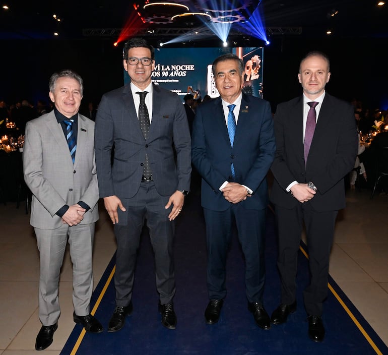 Cuatro hombres alineados en el evento, vistiendo trajes formales, con expresión tranquila y ambiente festivo.