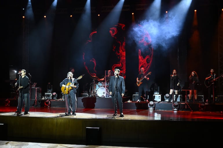 Seis músicos en escenario iluminado, guitarristas y vocalistas en acción. Ambientación musical con luces dramáticas y humo.