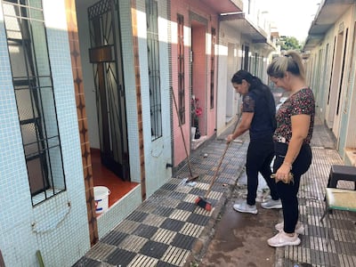 Dos mujeres en el pasillo de un edificio. Una limpia con escoba, la otra sonríe sosteniendo un objeto, rodeadas de puertas color pastel.