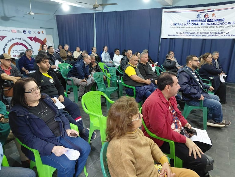 Grupo de personas escuchando atentamente en una sala de conferencias, con chaqueta oscura y camiseta amarilla al frente.
