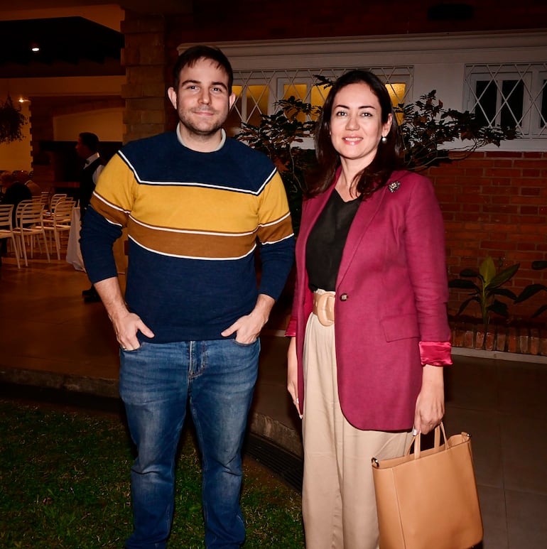 Mujer sonriente con blazer burdeos y bolso marrón, junto a hombre en suéter a rayas, en un ambiente social acogedor.