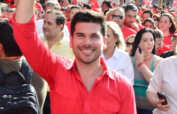 Hombre en camisa roja levantando su mano, rodeado de un grupo diverso de personas, todos con sonrisas en un ambiente de celebración.