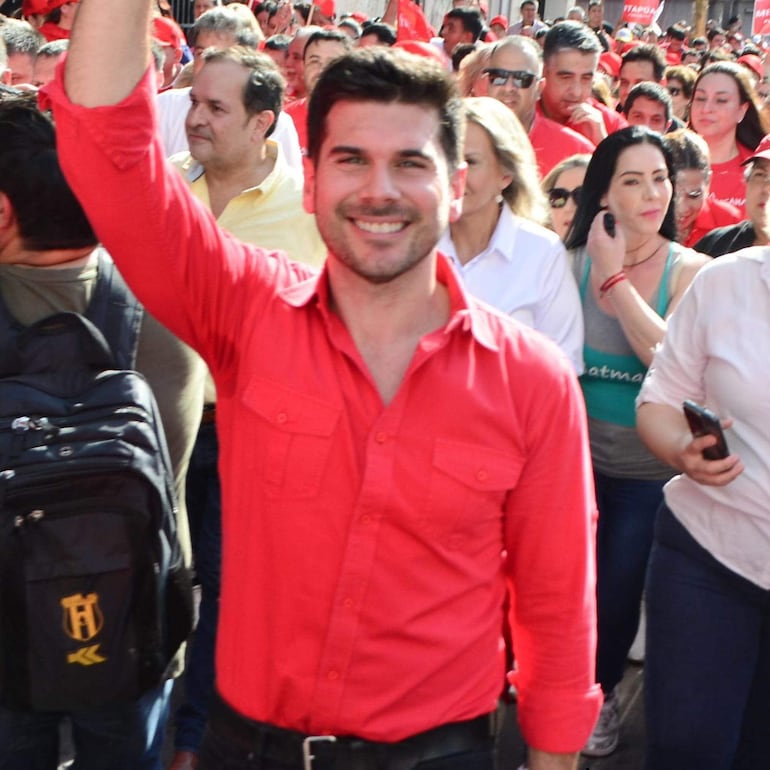 Hombre en camisa roja levantando su mano, rodeado de un grupo diverso de personas, todos con sonrisas en un ambiente de celebración.