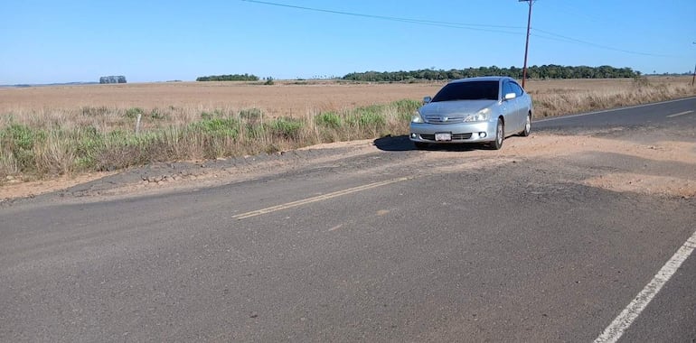 Un vehículo plateado estacionado junto a un camino dañado, rodeado de terreno abierto y árboles dispersos en un ambiente rural.