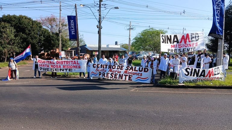 Grupo de médicos con prendas blancas sosteniendo pancartas sobre salud, en un ambiente exterior con cielo claro.