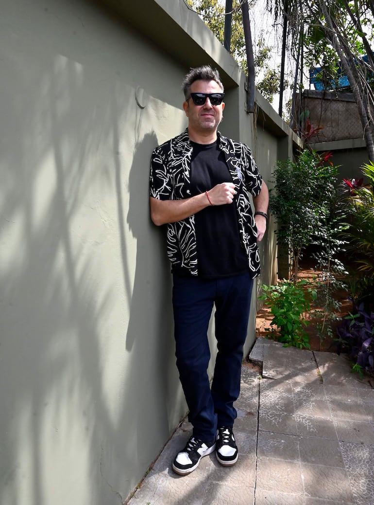 Hombre canoso con camisa negra y pantalones oscuros, apoyado en una pared verde entre plantas.