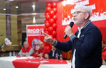Hombre canoso y gafas en chaqueta azul habla con pasión frente a asistentes con camisetas rojas en un evento decorado con globos.