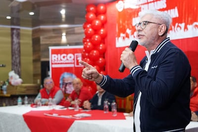 Hombre canoso y gafas en chaqueta azul habla con pasión frente a asistentes con camisetas rojas en un evento decorado con globos.