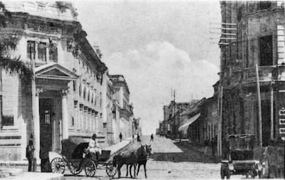 Dos personas en una calle de Asunción; una viste ropa tradicional y otra conduce un carro tirado por caballo entre edificios coloniales.