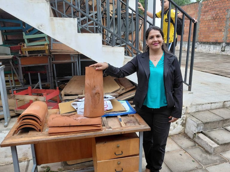 Mujer sonriente con blusa turquesa y pantalón negro sostiene cerámica, rodeada de documentos y un niño con camiseta amarilla al fondo.