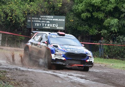 Automóvil de rally avanzando rápido por camino de tierra, levantando barro y agua, en un entorno natural con árboles.