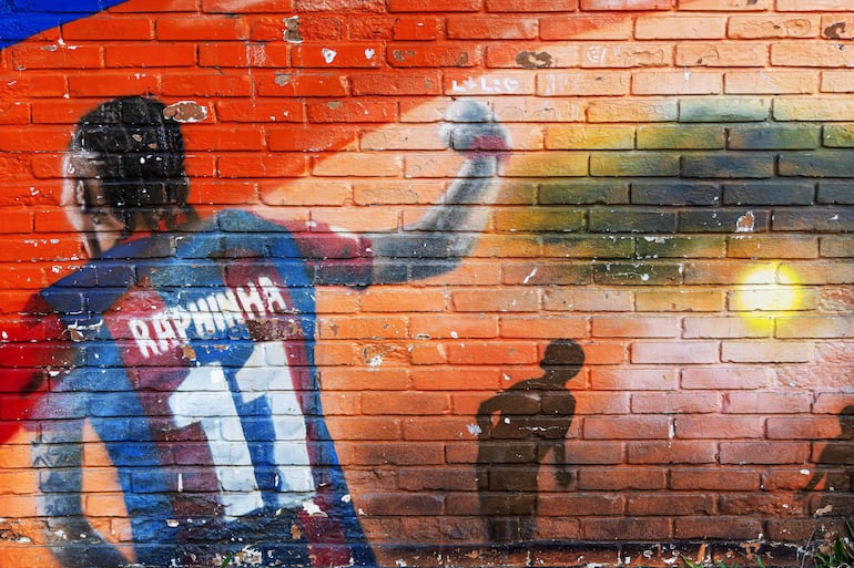 TOPSHOT - The shadow of a youngster is cast on a mural depicting Brazilian international football player Raphinha past a wall of the Engineer Ildo Meneghetti school in the Restinga neighborhood of Porto Alegre, Rio Grande do Sul state, Brazil, on May 16, 2026. Raphinha studied at the school during his childhood. (Photo by SILVIO AVILA / AFP)