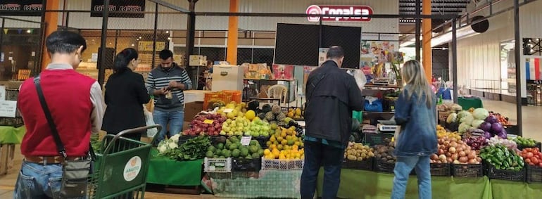 Hombre de espaldas con chaleco rojo, mujer en negro mirando su teléfono, varios compradores entre cajas de frutas y verduras.
