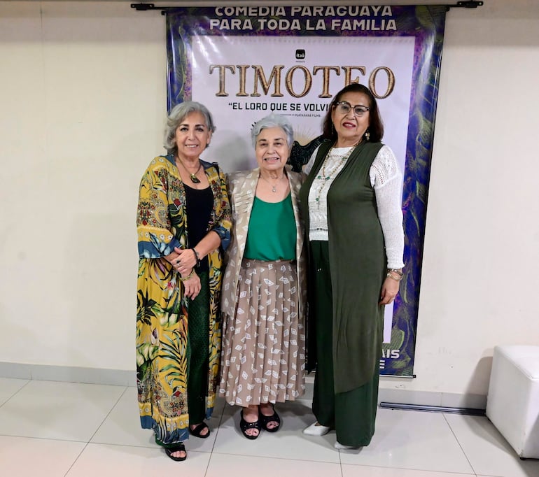 Tres mujeres de cabello canoso sonríen. Visten atuendos variados frente al cartel de 'Timoteo - El loro que se volvió'.