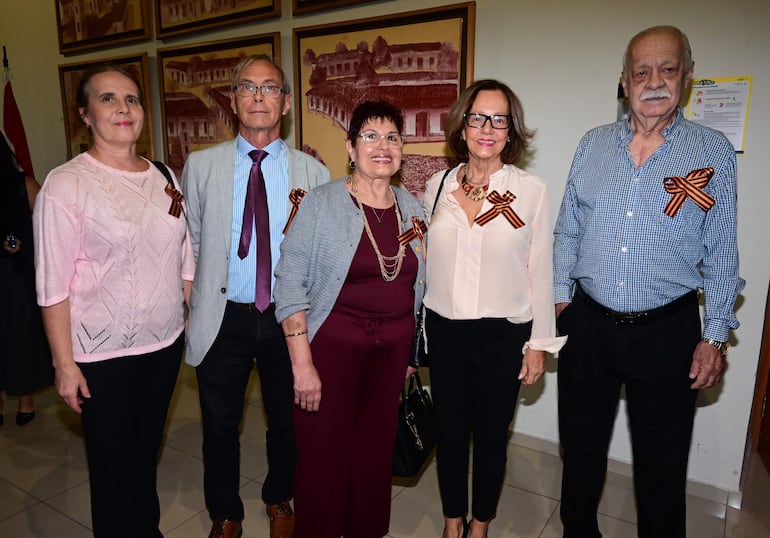 Cinco personas, cuatro mujeres y un hombre, de pie con cintas naranjas, en un ambiente formal. Fondo decorado.