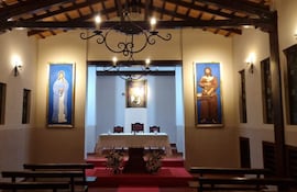 Interior de la capilla decorada con alfombra roja, altar blanco y arreglos florales, en un ambiente tranquilo con luz cálida.