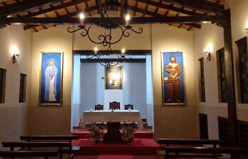 Interior de la capilla decorada con alfombra roja, altar blanco y arreglos florales, en un ambiente tranquilo con luz cálida.