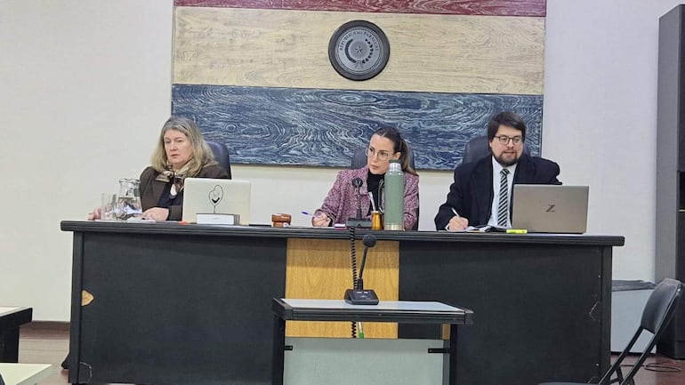 Tres personas en un tribunal: mujer con abrigo claro central, dos asistentes tomando notas en un ambiente formal. Bandera paraguaya en fondo.