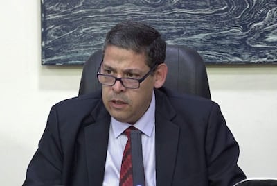 Hombre sentado con saco negro y corbata roja, gafas, expresión concentrada en un entorno de oficina.