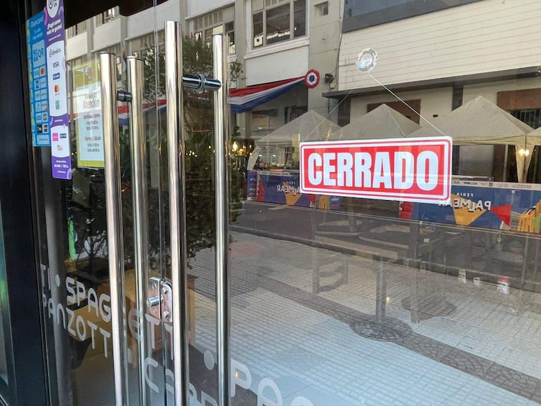 Letrero rojo que dice 'CERRADO' en puerta de cristal, con carteles visibles en un entorno urbano.