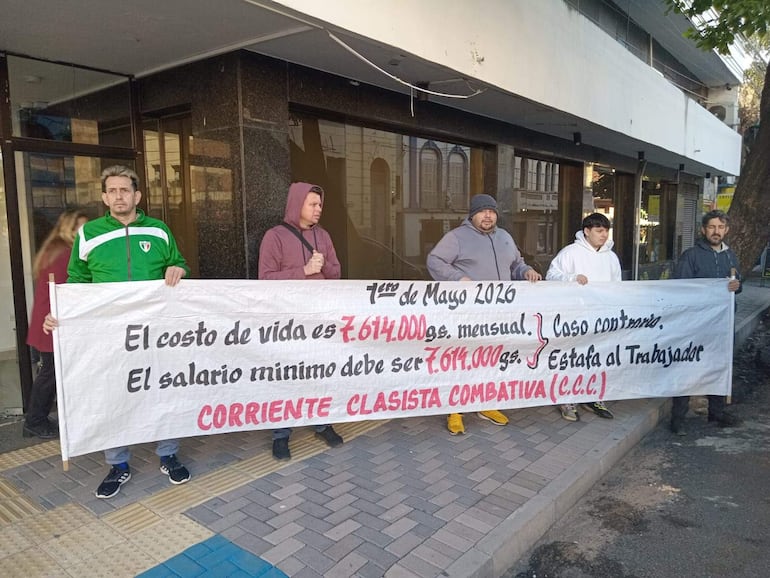 Cinco hombres en la acera, sosteniendo un gran cartel sobre salario mínimo. Uno lleva buzo verde y otro abrigo morado.
