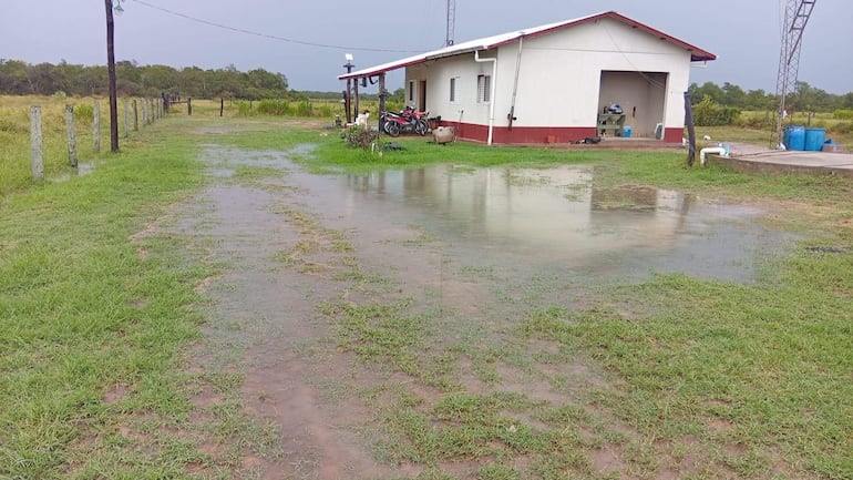 Entorno rural inundado con vivienda y motocicletas estacionadas, cielo nublado y cercas de cemento delimitando el área.