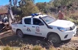 Vehículo policial blanco con logo de ITAIPU y retroexcavadora trabajando en terreno de vegetación, con operarios supervisando.