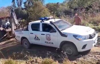Vehículo policial blanco con logo de ITAIPU y retroexcavadora trabajando en terreno de vegetación, con operarios supervisando.