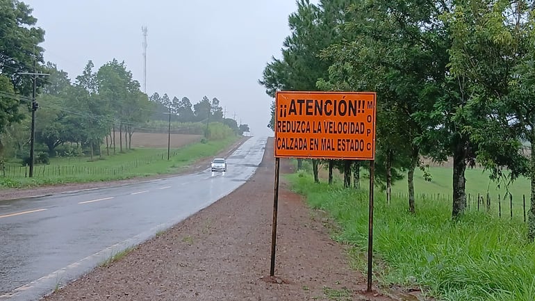 Cartel de advertencia naranja en carretera lluviosa, un coche circula bajo un entorno rural con vegetación abundante.
