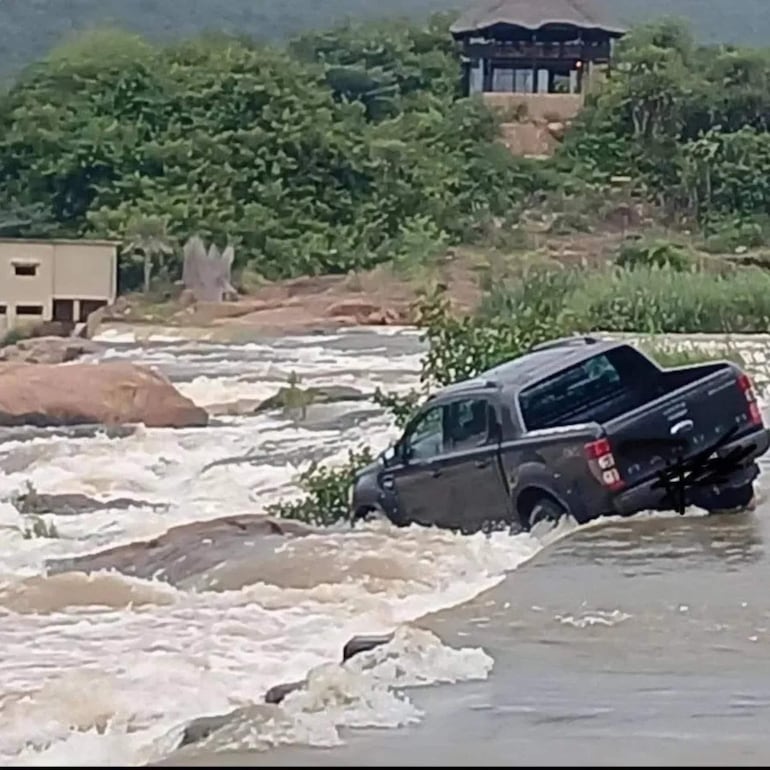 Camioneta negra parcialmente sumergida en agua, rodeada de árboles y un edificio de fondo, sin personas visibles.