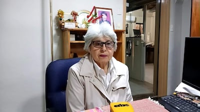 Mujer mayor con gafas y abrigo claro, sentada en escritorio con expresión neutral en oficina decorada con un pequeño altar.