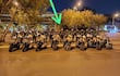 Grupo de 15 personas con uniformes negros alineados frente a motocicletas, observando atentamente en un entorno urbano con luces de hotel al fondo.