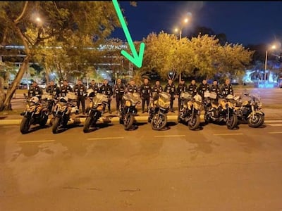 Grupo de 15 personas con uniformes negros alineados frente a motocicletas, observando atentamente en un entorno urbano con luces de hotel al fondo.