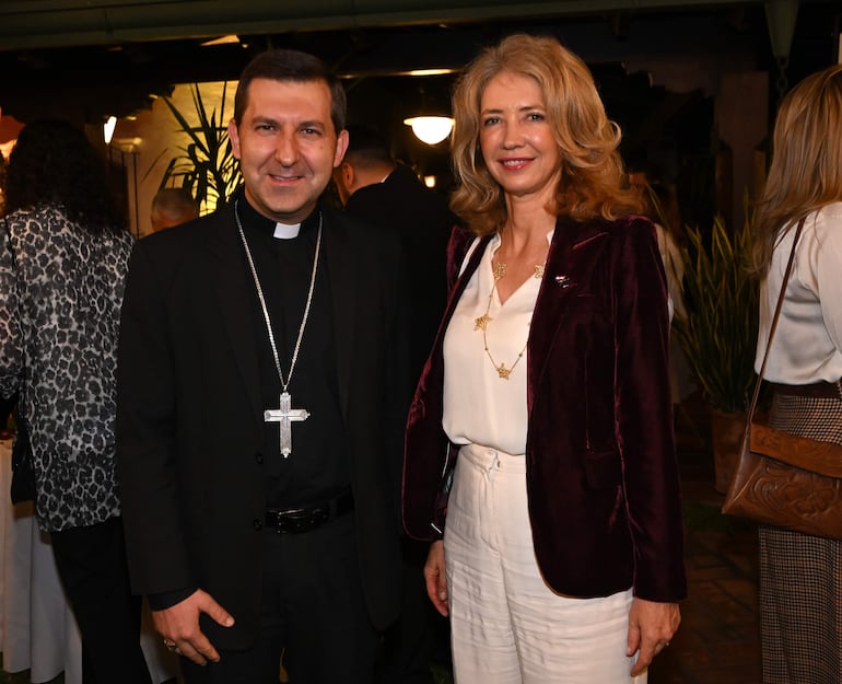 Hombre en vestimenta clerical y mujer en chaqueta de terciopelo sonríen en evento social decorado con vegetación.