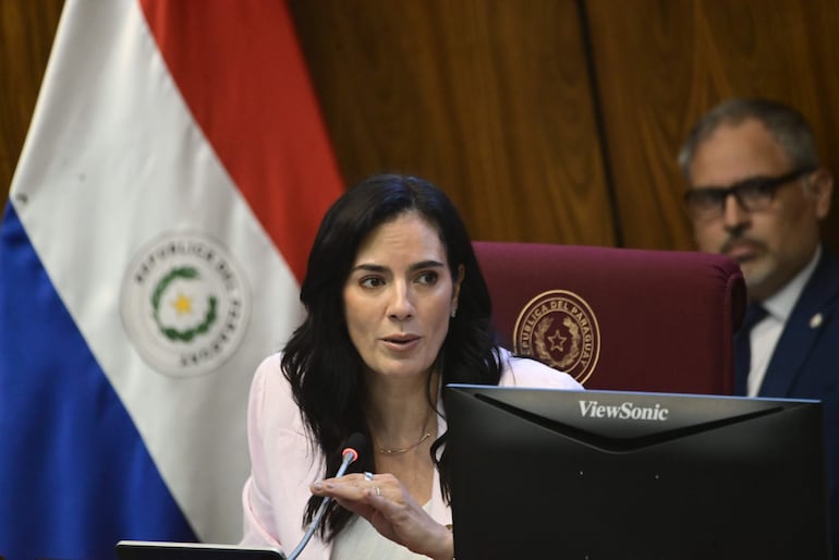 Claudia Centurión en chaqueta clara, hablando ante un micrófono, con banderas de Paraguay y un hombre atento al fondo.