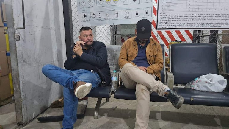 Dos hombres en espera en un ambiente interno; uno tocándose la barba y el otro mirando hacia abajo con concentración.