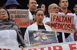 Mujer seria en el centro sostiene un cartel que dice 'EN MEMORIA DE LA DRA. GIGOSA BOGARÍN' junto a otros médicos con batas blancas.