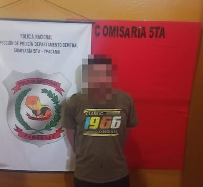 Hombre de 32 años en camiseta gris frente a cartel de la Comisaría 5ta de Policía Nacional en fondo naranja.