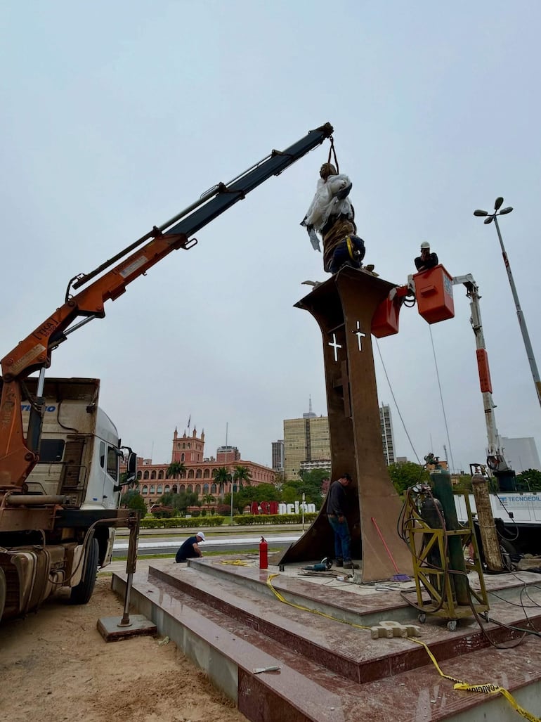 Cuatro trabajadores en grúa y en el suelo manipulan una figura cubierta mientras edificios históricos se ven al fondo.