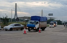 Vehicles en movimiento, incluyendo camiones y un automóvil gris, en una carretera con cielo nublado; Puente de la Amistad al fondo.
