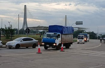 Vehicles en movimiento, incluyendo camiones y un automóvil gris, en una carretera con cielo nublado; Puente de la Amistad al fondo.