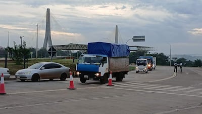 Vehicles en movimiento, incluyendo camiones y un automóvil gris, en una carretera con cielo nublado; Puente de la Amistad al fondo.