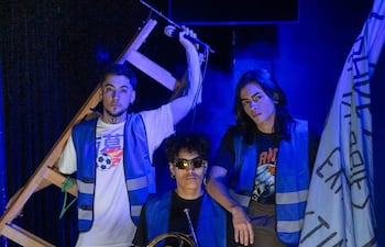 Tres músicos en escenario iluminado: uno con estructura de madera, otro sosteniendo un instrumento de viento y un tercero con bandera celeste.
