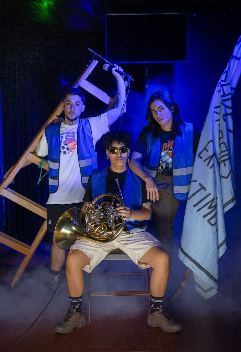 Tres músicos en escenario iluminado: uno con estructura de madera, otro sosteniendo un instrumento de viento y un tercero con bandera celeste.