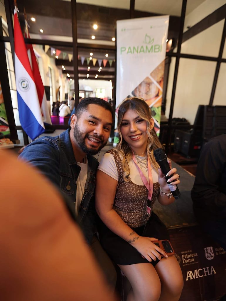 Amyris Díaz con micrófono y Oliver Ferreira sonriendo, rodeados de banderas multicolores y un público en un ambiente festivo.