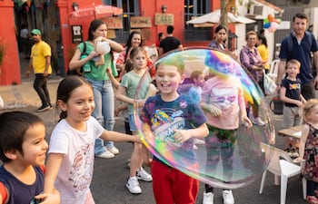 Niños sonrientes con camisetas coloridas, atrapando burbujas en un entorno festivo con adultos observando en un café colorido.